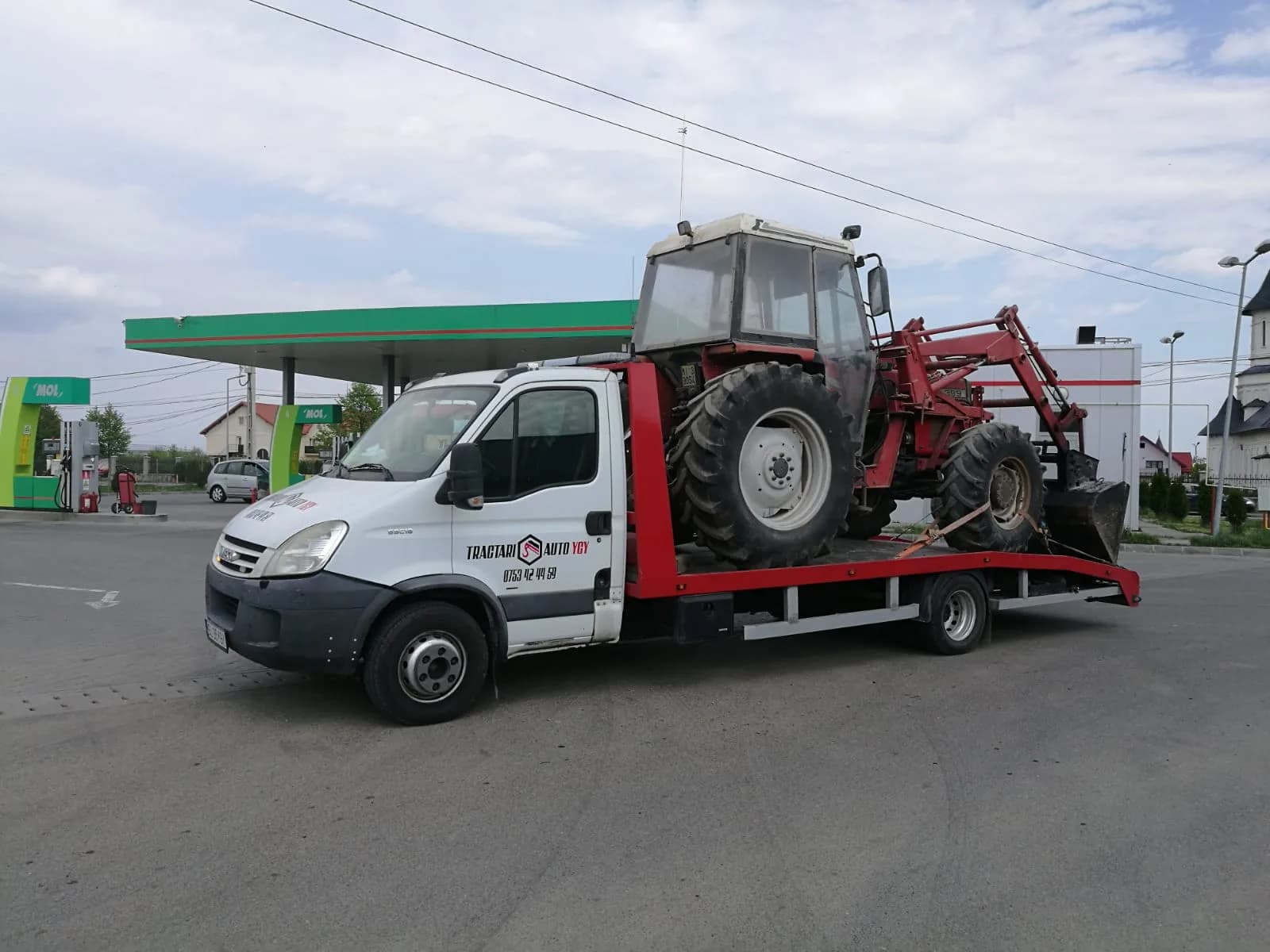Tractor agricol transportat pe platformă, oprire într-o benzinărie.